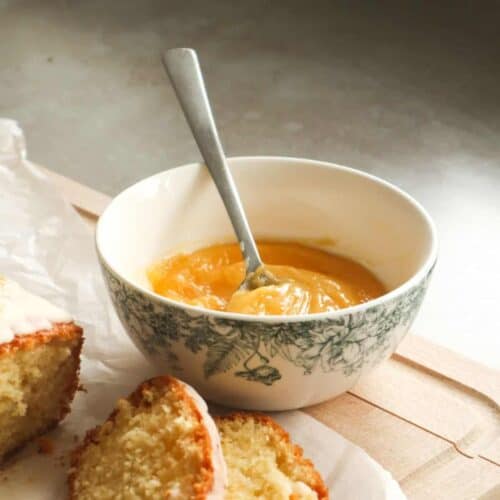 Een close-up van zelfgemaakte lemon curd in een wit schaaltje op een plank met wat cake en bakpapier.