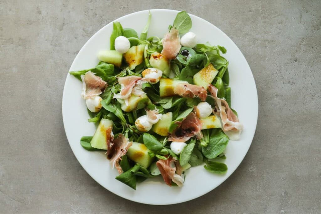 een salade op een wit bord met mozzarella, parmaham, meloen en veldsla